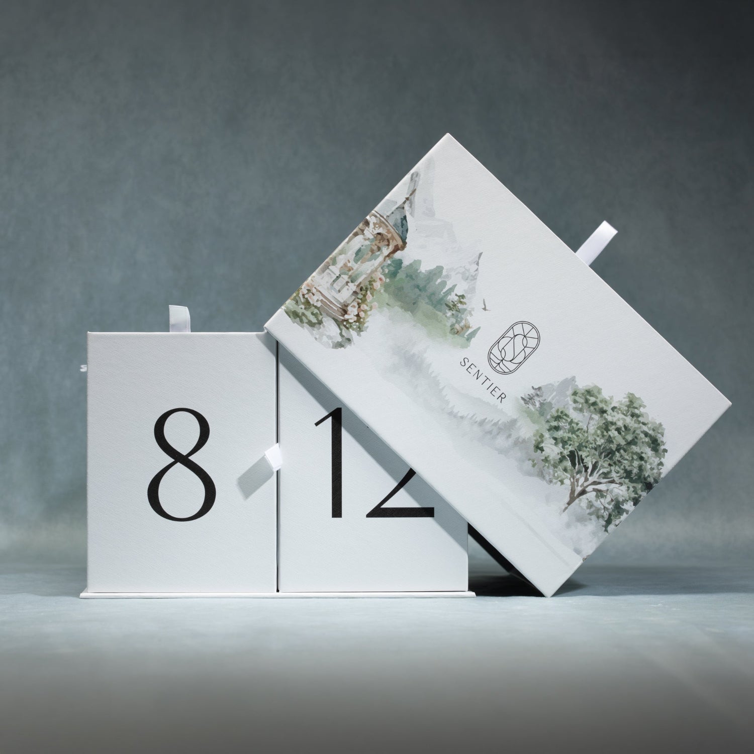 A white calendar-style box set with large black numbers on the front panels and a lid decorated with soft green and gray watercolor trees and a small 'Scentier' logo, photographed on a gray studio background.